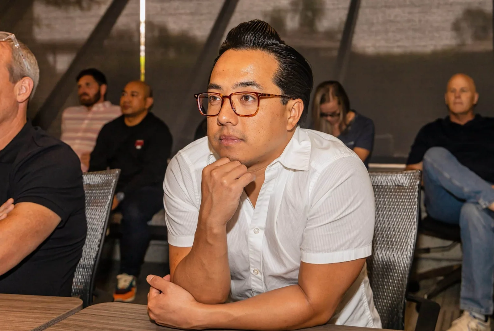 A LightBox colleague in a white shirt listening with his chin on his hand, at a long table of colleagues during a team session.