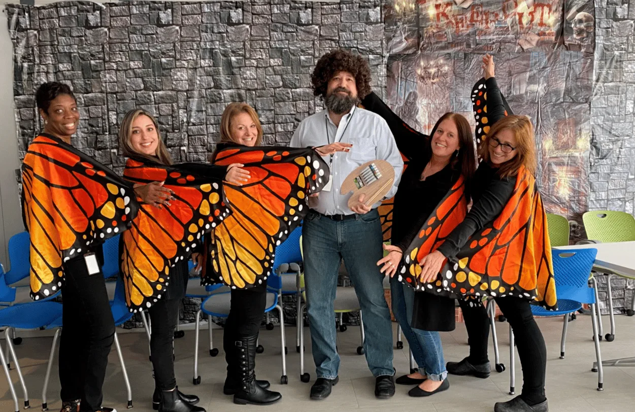 Six LightBox colleagues in an office meeting room wearing bright orange monarch butterfly wings, arms spread, on a costume day.